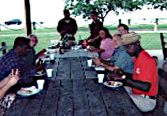 A number of people at a picnic table with others in the background.